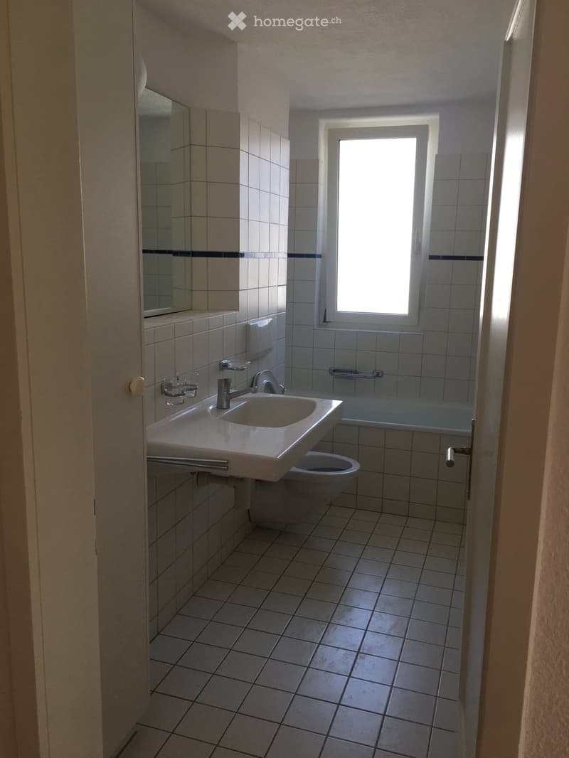 Bathroom with white tiled walls and floor, a sink with a faucet, a toilet, a bathtub, and a mirror above the sink. A window provides natural light.