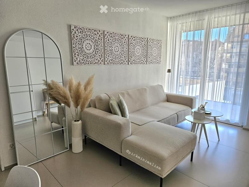 Modern living room with a light grey sectional sofa, ottoman, coffee table, side table, decorative wall panels, and a large mirror. The room features large windows with sheer curtains and a vase with dried pampas grass.