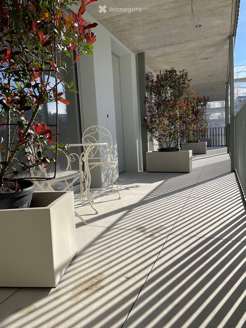 Modern balcony with white tiled floor, white metal chair and table, several potted plants, and a covered area with a concrete ceiling.