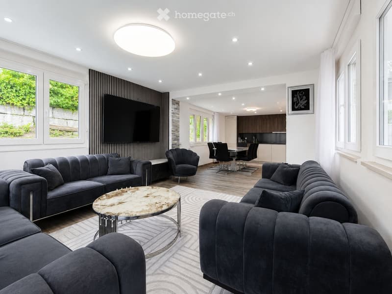 Modern living room with grey sectional sofa, coffee table, armchair, dining area with chairs and table, and a large television mounted on the wall. The space features large windows with a view of greenery outside.