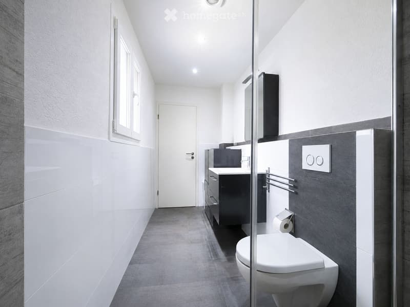 Modern bathroom with white tiles, grey accents, a toilet, sink, mirror, and towel rack.