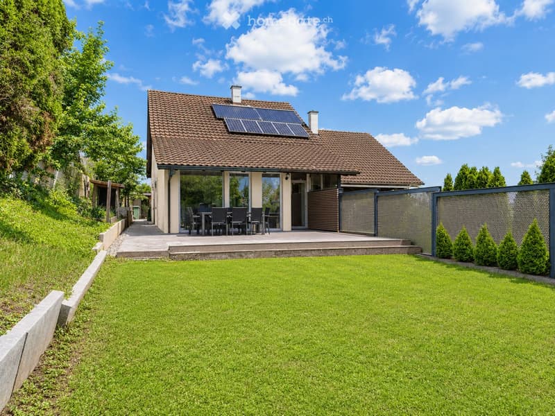 Two-story house with a tiled roof, solar panels, and a modern design. The house features a raised ground floor, a patio with outdoor furniture, and a well-maintained lawn.