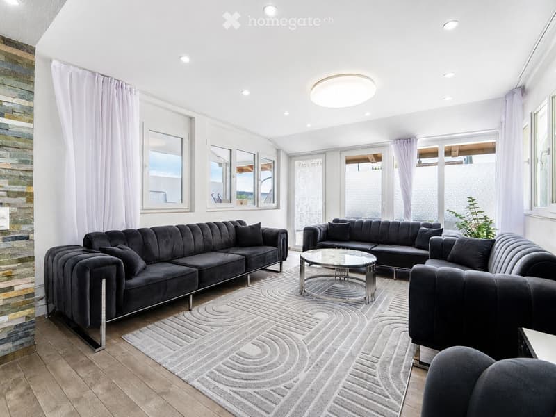 Modern living room with grey sectional sofa, coffee table, and armchair, large windows, and a view of the outdoor area.