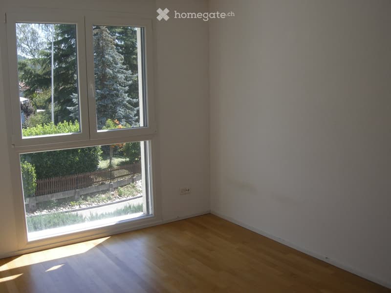 Empty room with light wooden flooring and large windows offering a view of a garden and trees.
