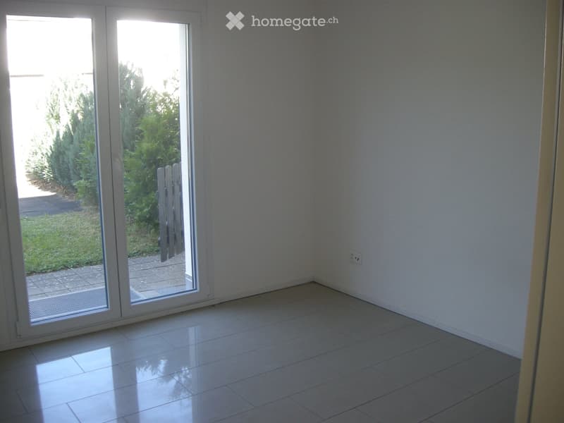 Empty room with white tiled flooring and white walls, featuring a large sliding glass door leading to a garden area.