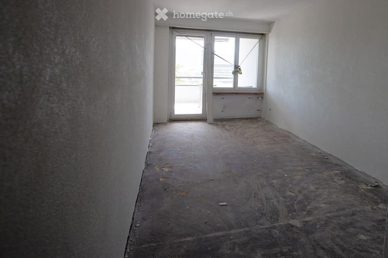 Empty room with bare walls, concrete floor, and a large window with a sliding door leading to a balcony.