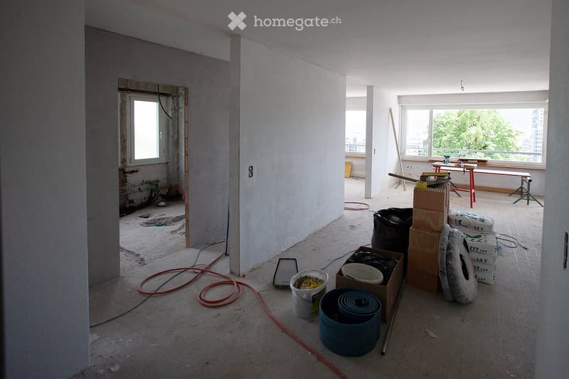 Empty room with unfinished walls, exposed pipes, and construction materials scattered around. Windows provide natural light, and the floor is covered in dust and debris.
