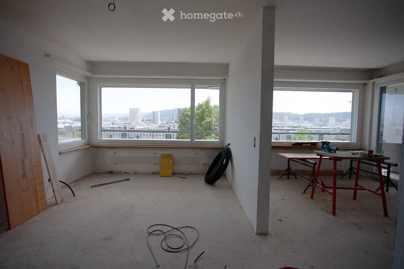 Empty room with large windows offering a cityscape view, exposed wooden planks, and construction materials scattered around.