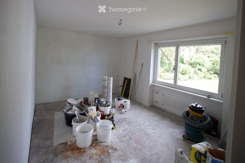 Empty room with bare walls and unfinished floor, containing construction materials and tools, including buckets, a vacuum cleaner, and a window with a view of greenery.
