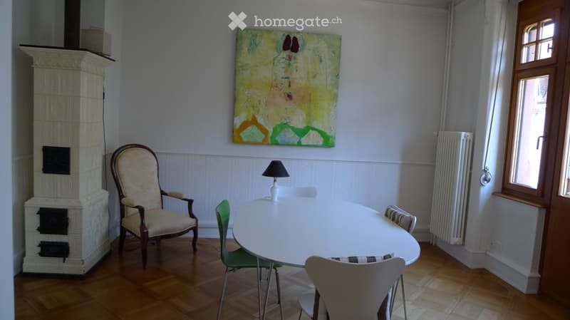 Dining area with a round white table, four chairs, a lamp, a radiator, and a colorful painting on the wall.