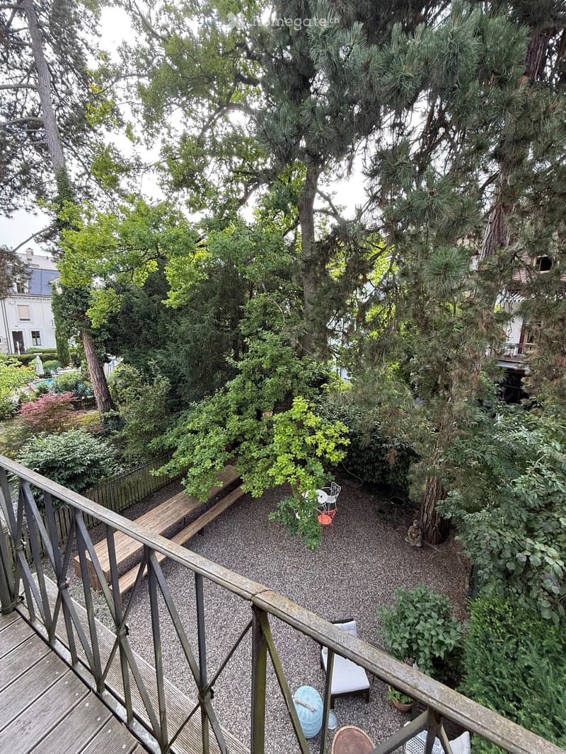 Garden area with gravel ground, wooden bench, small seating area with chairs and table, surrounded by trees and greenery.
