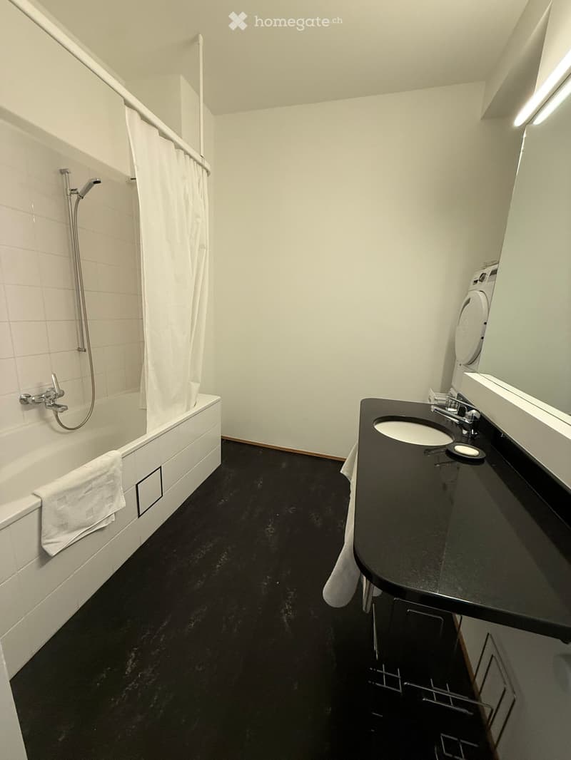Modern bathroom with a black countertop sink, white bathtub, and a shower area with a curtain. The floor is black, and there is a towel on the edge of the bathtub.