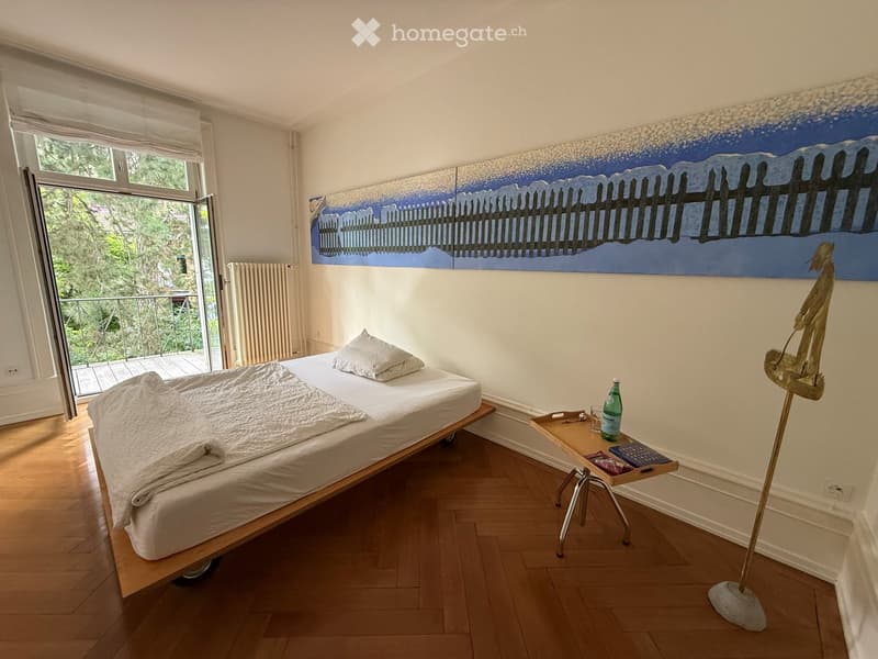 Cozy bedroom with a wooden bed, nightstand, and large painting on the wall. Features a balcony with outdoor view.