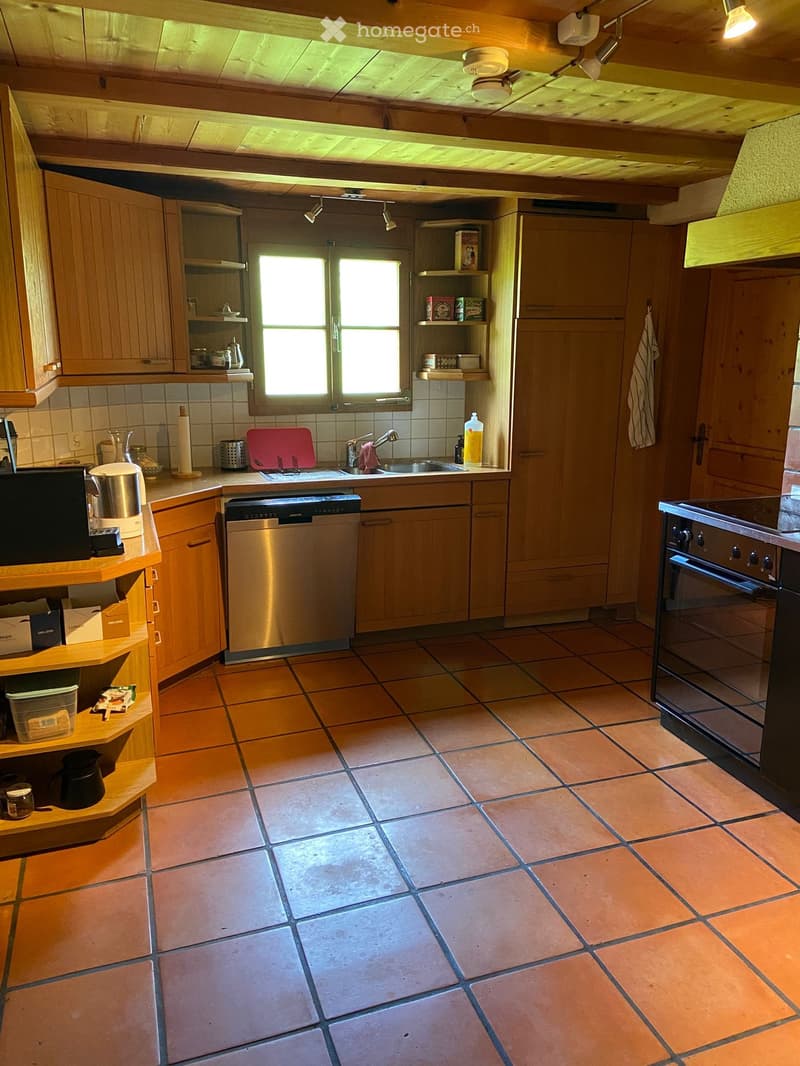The kitchen features wooden cabinets, a tiled floor, and stainless steel appliances including a sink, oven, and dishwasher. There is a window above the sink, and various kitchen items are placed on the countertops and shelves.