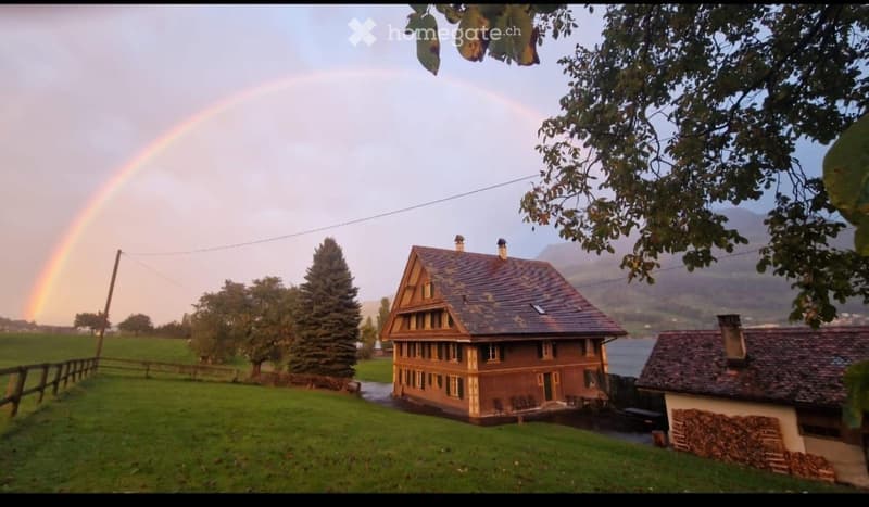 A two-story wooden house with a dark roof, surrounded by a green lawn and trees, with a rainbow in the sky and a view of a lake and mountains in the background.