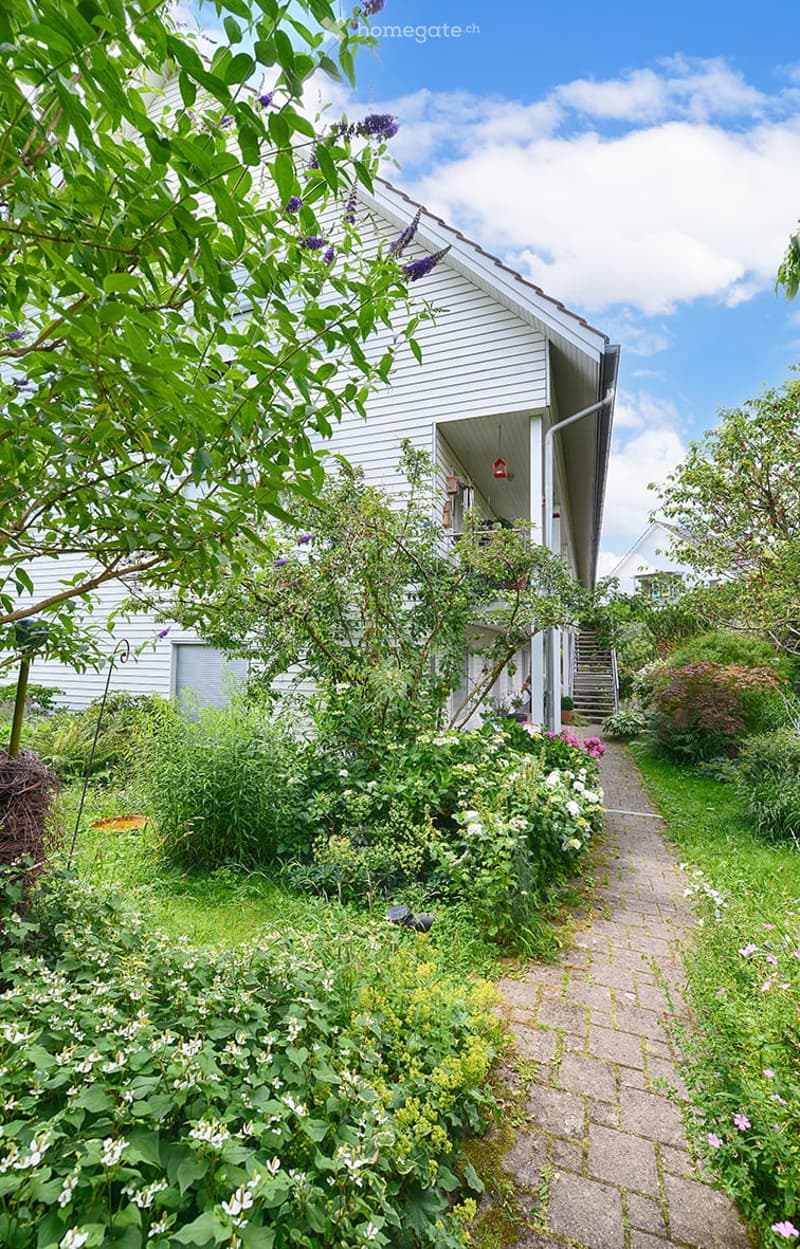 Two story house, white walls, wooden balcony, raised ground floor, brick pathway, plants, trees