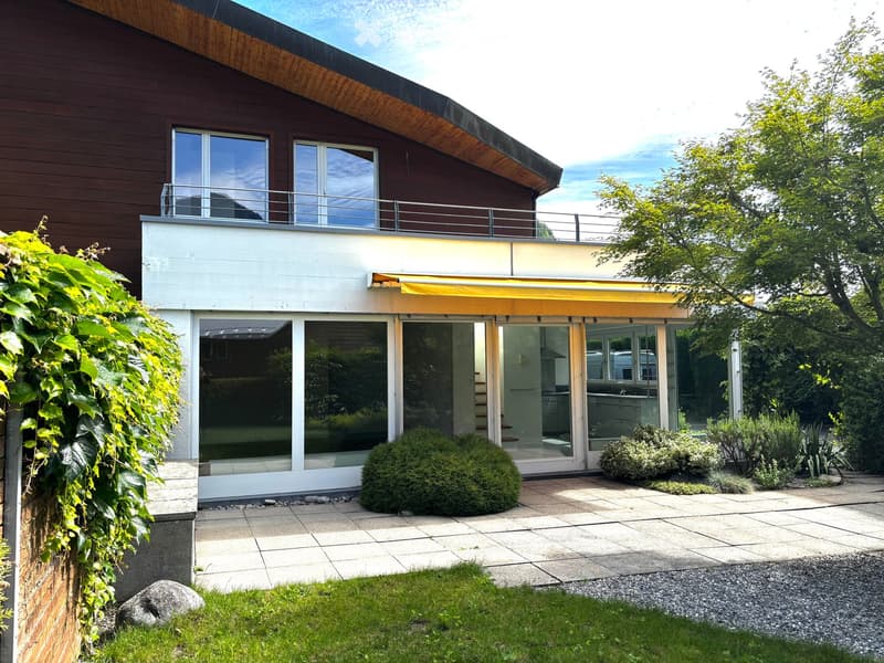 Modern two-story house with large windows, a balcony, and a well-maintained garden area with a patio and greenery.
