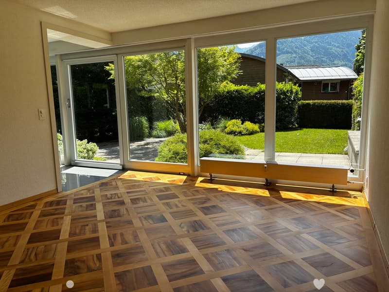 Empty room with wooden herringbone floor, large glass sliding doors opening to a garden, radiator, and a view of mountains and greenery.