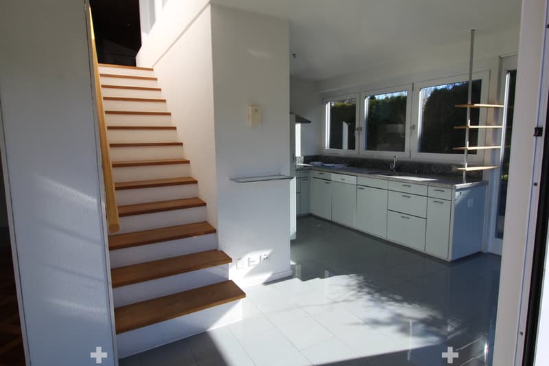 Bright, modern kitchen with white cabinets, a granite countertop, and a sink. A wooden staircase leads upwards, and large windows allow natural light to fill the space.