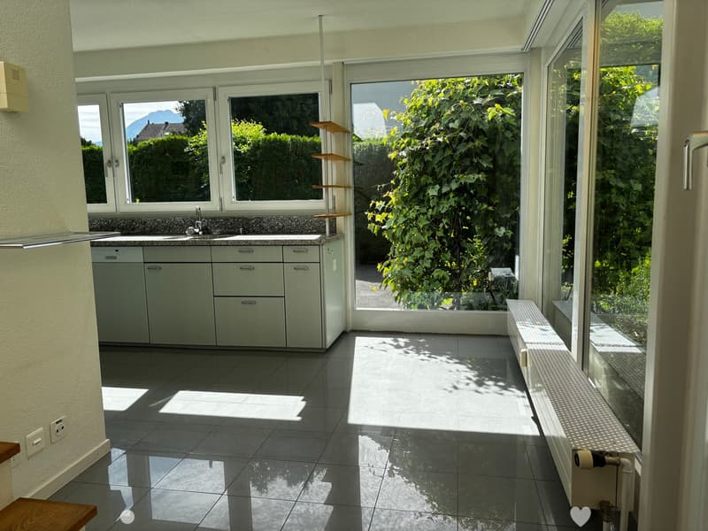 Bright, modern kitchen with large windows, light grey cabinets, a sink, and a dishwasher. The floor is tiled, and there is a small shelf unit near the window.