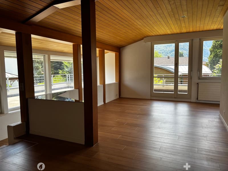 Bright, unfurnished room with wooden flooring and wooden ceiling beams, large windows and sliding doors leading to a balcony with a scenic mountain view.