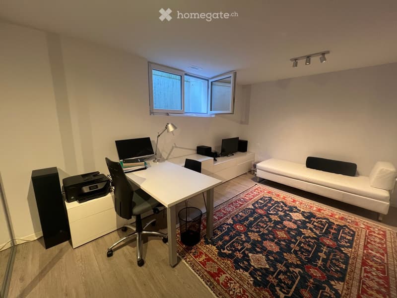A modern, multi-functional room with a white desk, ergonomic chair, laptop, printer, and a sleek white sofa. The room features a large window, a patterned rug, and built-in storage units.