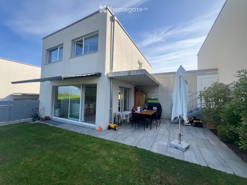Modern house with a well-maintained garden and patio area. The patio features a dining table, chairs, an umbrella, and a toy car. The house has large windows and a covered outdoor area.