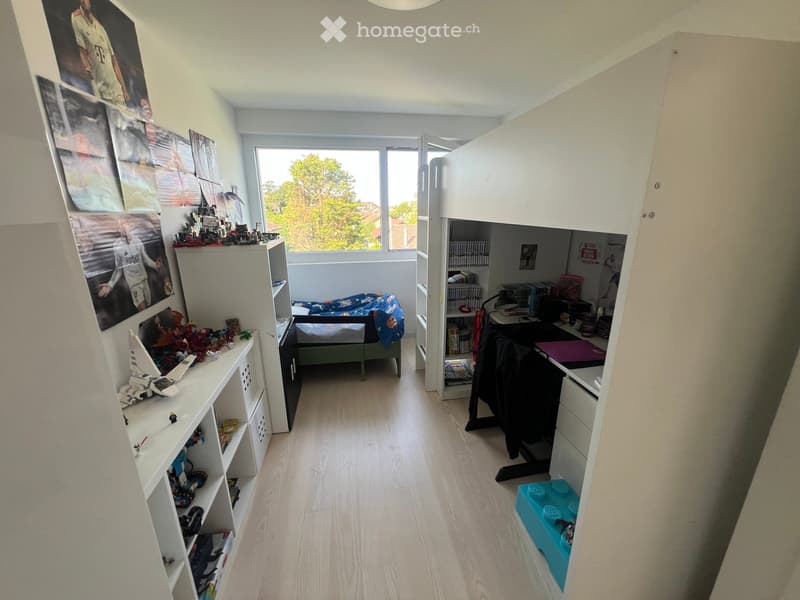 A well-lit bedroom with a bed, shelves filled with toys and books, a desk with a chair, and a large window offering a view of greenery outside.