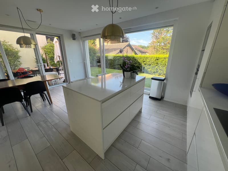Modern kitchen with a white island, pendant lights, and a dining area visible through large windows.