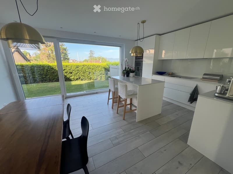 Modern kitchen with white cabinets, a central island, pendant lights, and a large window overlooking a garden.