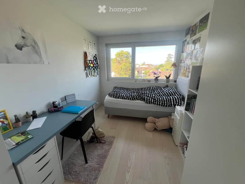 Cozy bedroom with a single bed, desk, chair, bookshelf, and small plants. The room features a large window with a view of trees and a cityscape.