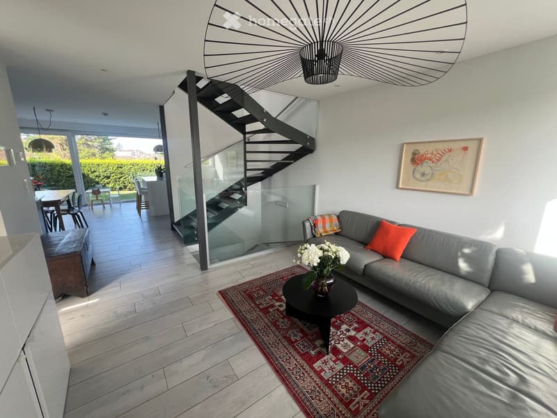 Modern living room with a grey sectional sofa, coffee table with a flower arrangement, and a staircase leading to an upper level. The room features a patterned rug, a glass railing, and a dining area visible in the background.