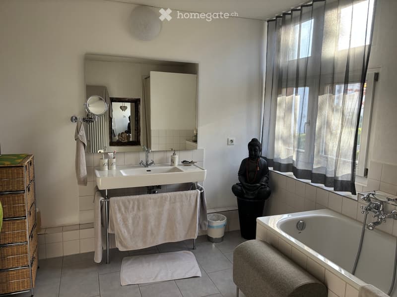 white bathtub, vanity with double sinks, large mirror, toilet, Buddha statue, small cabinet, shelves, curtained window, towel rack