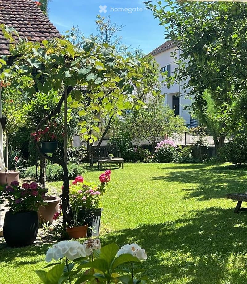 Garden with lawn, flowers, garden bench, and a small table. A house is in the background.