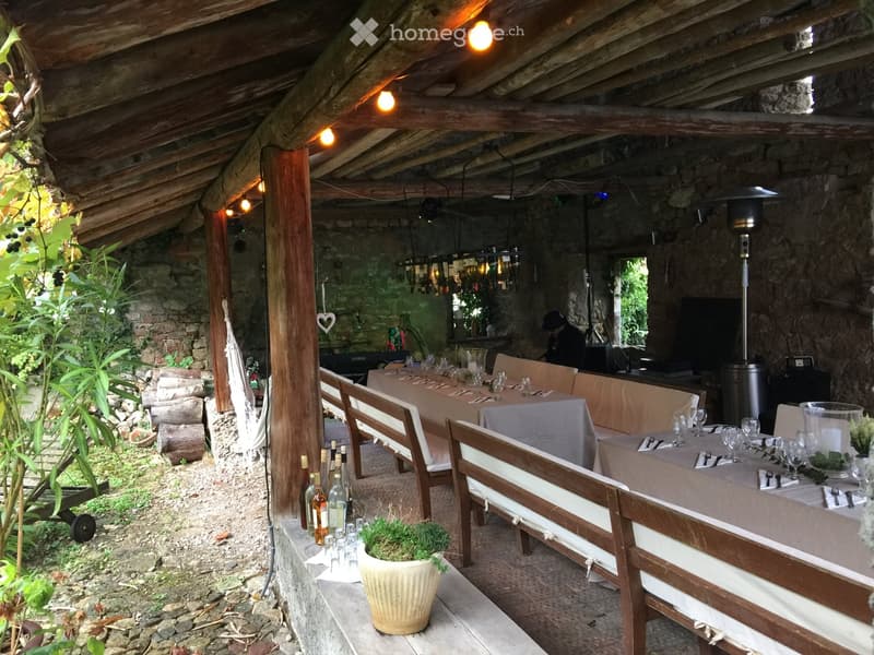 Outdoor dining area with wooden benches, long tables, hanging lights, plants, and bottles of wine.