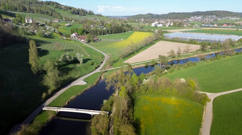 Aerial view of a rural landscape with fields, a river, a bridge, and scattered houses and trees.