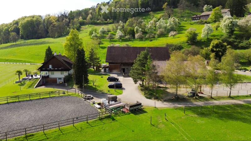 Aerial view of a rural property with multiple buildings, including a main house, a barn, and a smaller structure. The property features well-maintained lawns, a gravel driveway, fenced areas, and several parked cars. The surrounding landscape includes rol