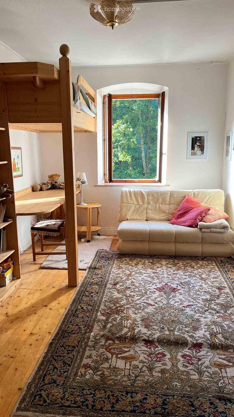 Cozy living room with a cream sofa, colorful pillows, large patterned rug, wooden floor, and a lofted bed area with a small table and chairs.