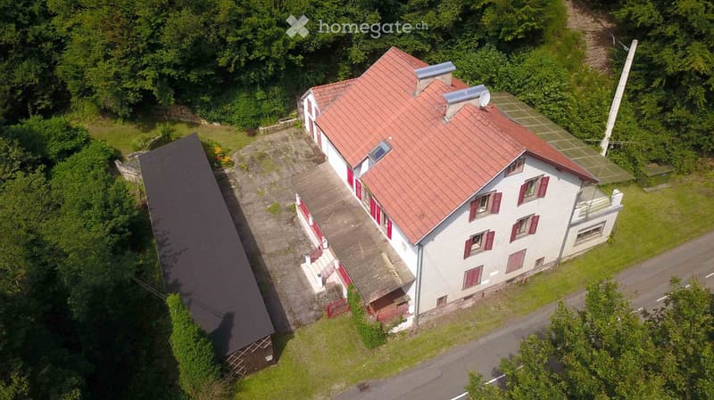 Aerial view of a two-story house with red-tiled roof, white walls, and red shutters. The house has a driveway, a small garage, and is surrounded by greenery.
