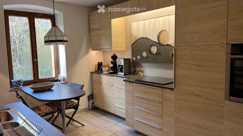 Modern kitchen with light wood cabinets, stainless steel appliances, a black countertop, and a small dining area with a table and chairs.