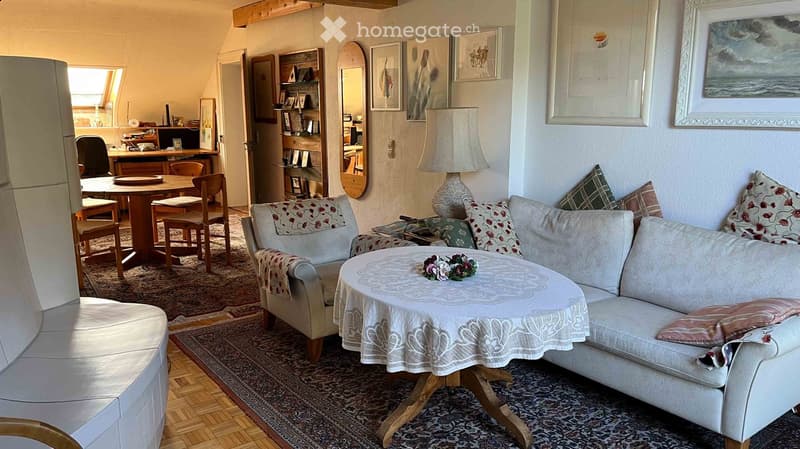 Cozy living room with a cream-colored sofa, coffee table, and wooden dining table with chairs in the background. The room features a patterned rug, framed artwork, and a small decorative centerpiece on the coffee table.