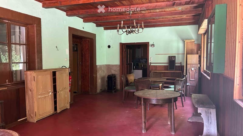 A rustic room with wooden furniture, including a central table, chairs, a bench, and a cabinet, featuring a chandelier and a red carpeted floor.