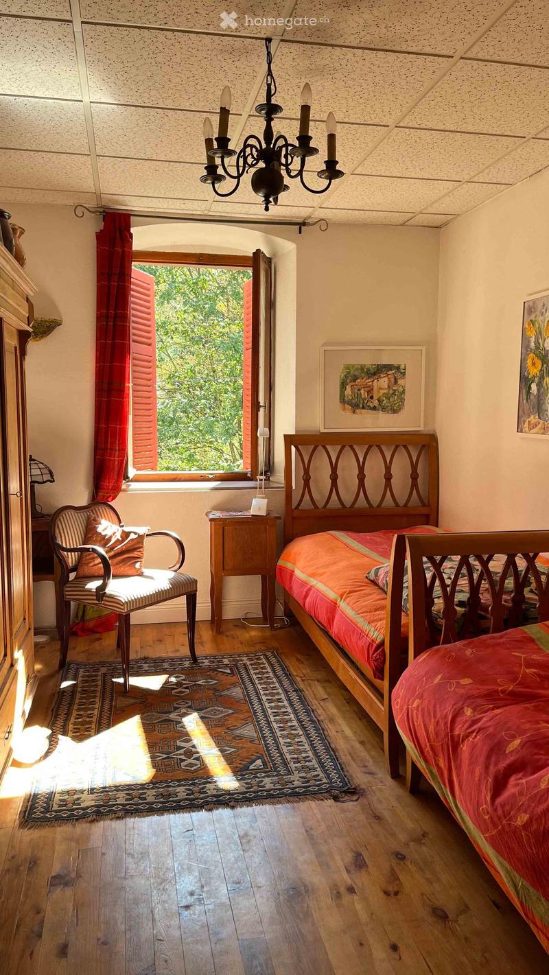 Cozy bedroom with wooden floors, a large bed with red bedding, a wooden chair with striped upholstery, a nightstand, and a chandelier hanging from the ceiling. A large window with red curtains offers a view of greenery outside.