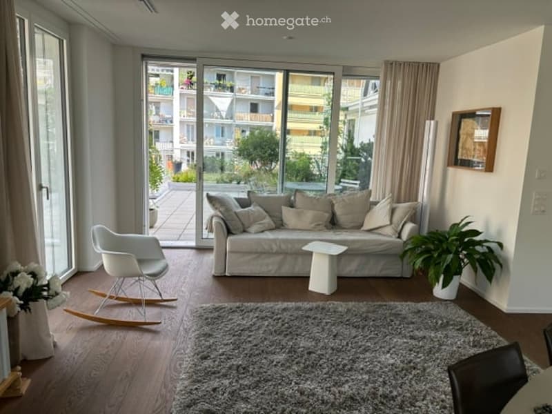 Modern living room with large windows and balcony access, featuring a white sofa, coffee table, white rocking chair, and a potted plant.