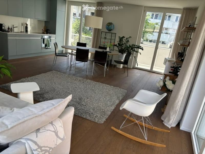 Open-plan living and dining area with modern furniture, large windows offering a view, a sofa, coffee table, rocking chair, dining table with chairs, kitchen area with cabinets and appliances, floor lamp, potted plant, and a wine rack.