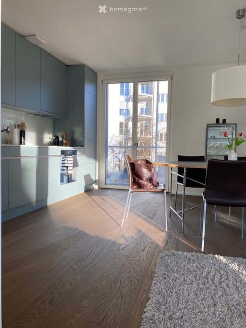 Modern kitchen and dining area with light blue cabinets, wooden flooring, and a large window with a view of the city.