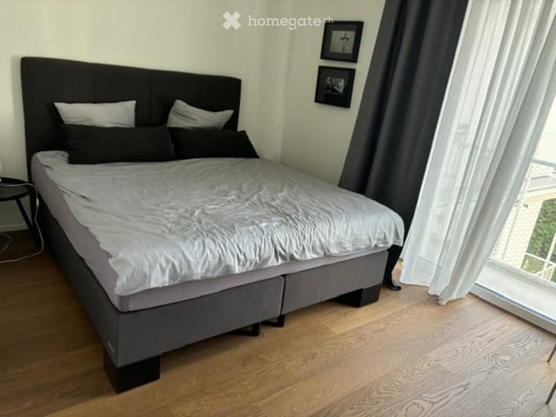 Modern bedroom with a large double bed, grey bedding, wooden floor, nightstand, and framed pictures on the wall. Large window offers a view.