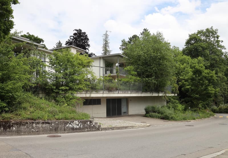 Modern two-story building with a raised ground floor, surrounded by lush greenery and trees. Features a balcony on the upper level and a paved driveway leading to the entrance.