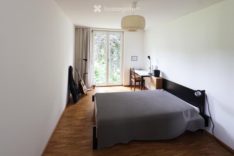 Bright bedroom with wooden flooring, large window with sliding doors leading to a balcony, bed with grey duvet, guitar and case, small desk with chair, and wall-mounted lamp.