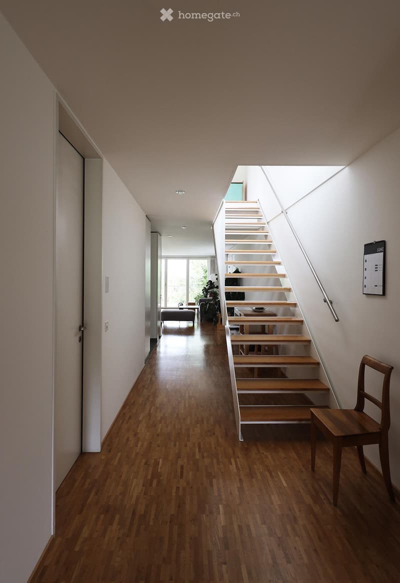 Modern hallway with wooden flooring, white walls, and a staircase leading upwards. Features a small seating area with a chair and table, and a large window at the end.